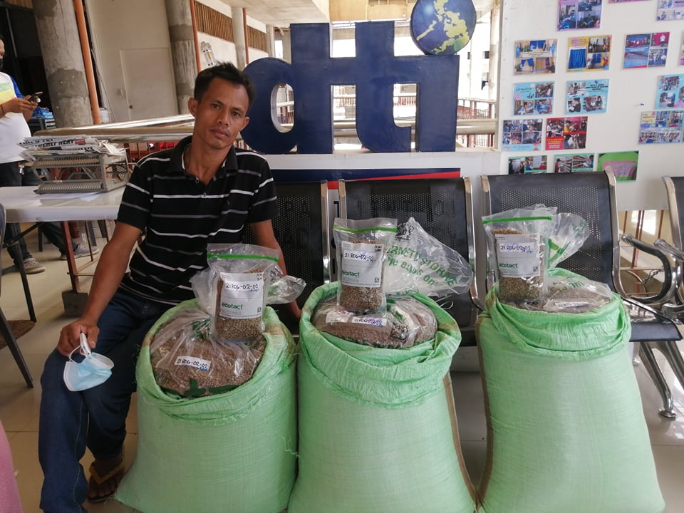 A man poses beside three sacks of coffee beans, a signage bearing the DTI logo behind him